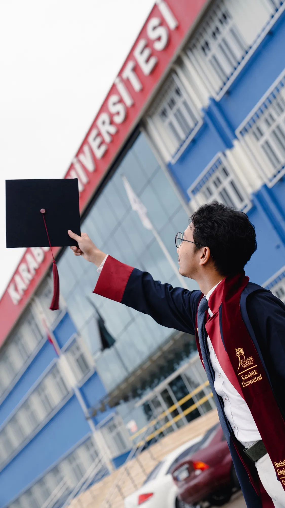 Hasbii in a graduation gown, holding a cap up outside Karabük University