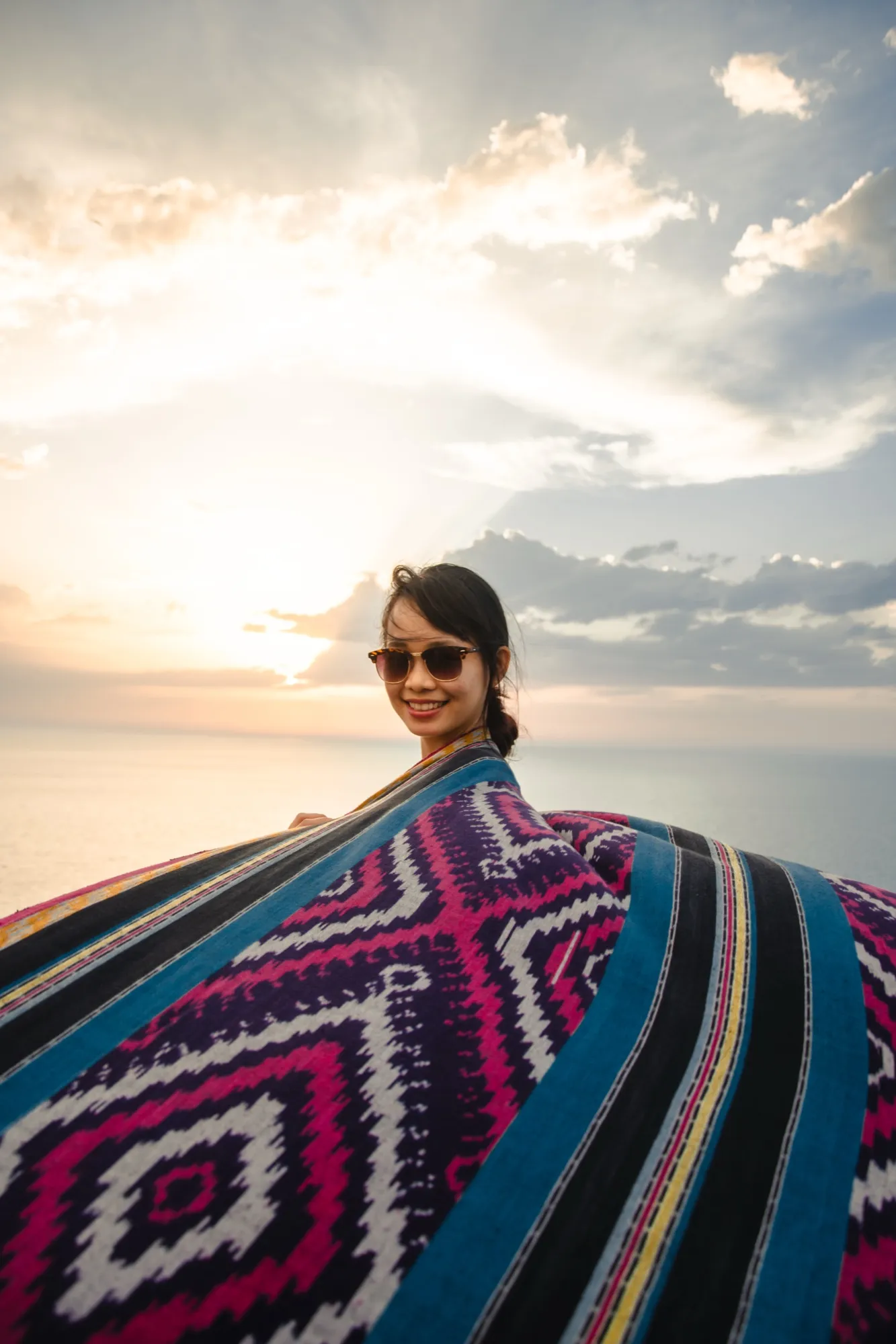 A woman in sunglasses holding a woven blanket open to the wind at sunset