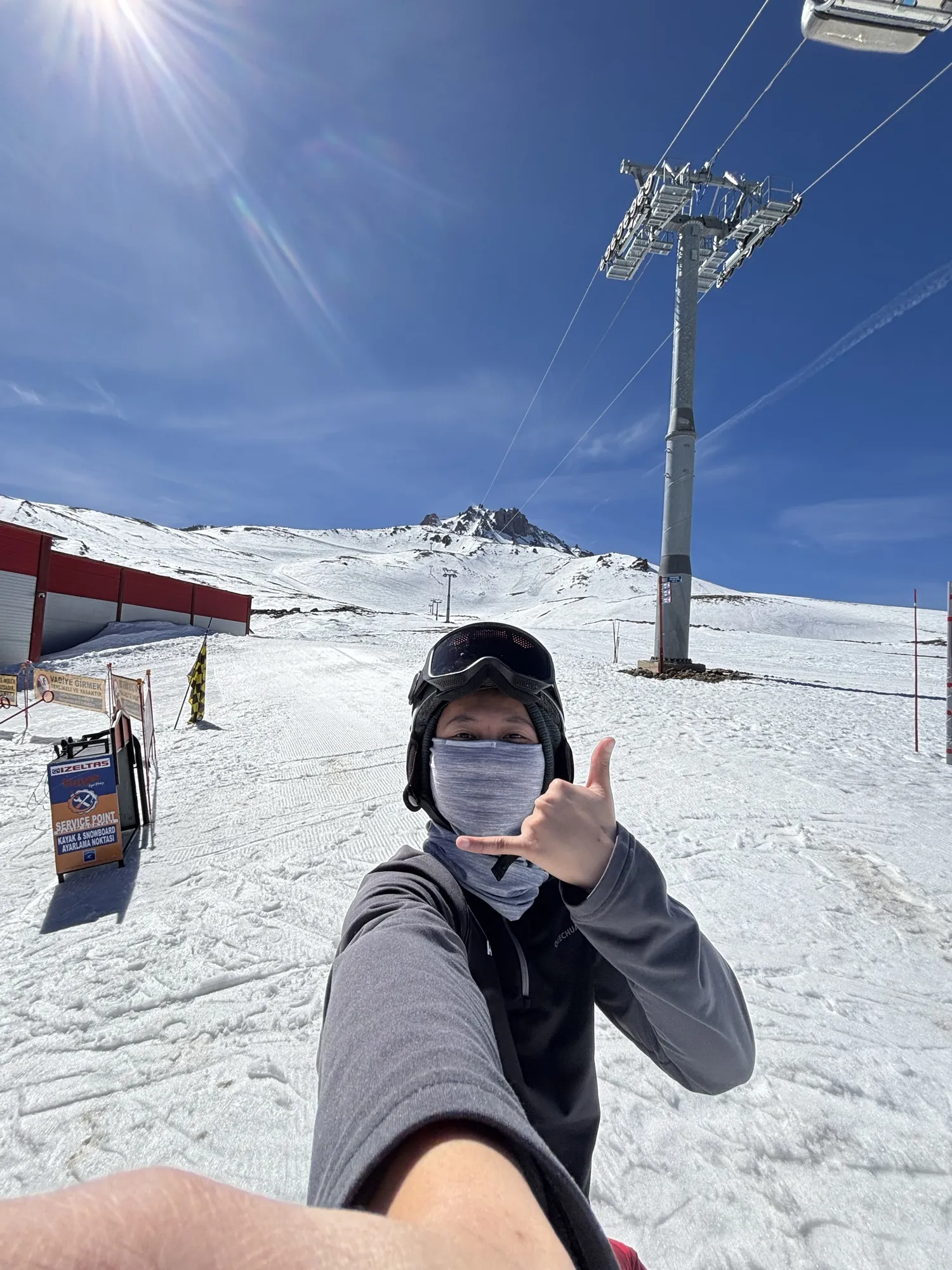Hasbii in goggles and a face mask throwing a hang-loose sign under a ski lift